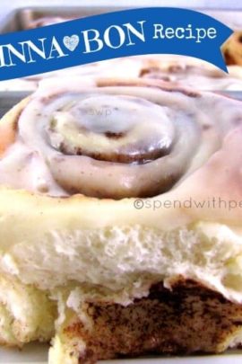 single copy cat cinnabon with frosting on a white plate with a tray of cinnamon buns in the background