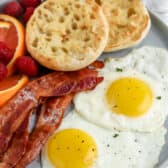 two fried eggs on plate with english muffin, bacon, and fruit