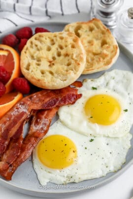 two fried eggs on plate with english muffin, bacon, and fruit